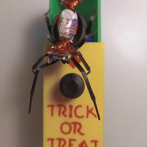 Puede incluir: Decoración de Halloween con una araña hecha de envoltorios de caramelos sobre una caja amarilla con la inscripción "TRICK OR TREAT" en rojo. Detrás de la araña hay una caja verde.
