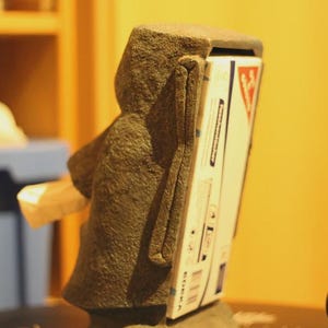 Pu&ograve; includere: Una scultura in pietra a forma di testa Moai, con una scatola bianca e rossa inserita sul retro. La scatola presenta testo e grafica. La scultura &egrave; di colore grigio scuro.