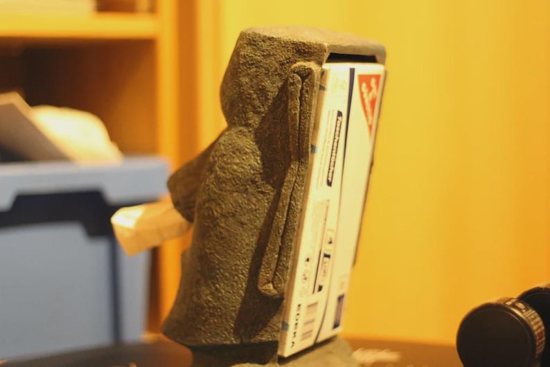 Pu&ograve; includere: Una scultura in pietra a forma di testa Moai, con una scatola bianca e rossa inserita sul retro. La scatola presenta testo e grafica. La scultura &egrave; di colore grigio scuro.