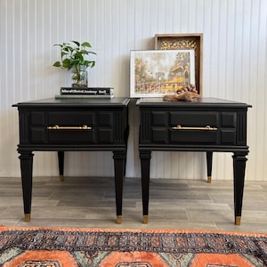 May include: Two black end tables with gold hardware and a single drawer. The tables are on a patterned rug with a white wall in the background.
