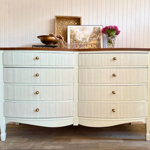 May include: A white dresser with six drawers and a dark wood top. The drawers have vertical striped detailing and brass knobs. The dresser is decorated with a framed picture, a decorative bowl, and a vase of pink flowers.