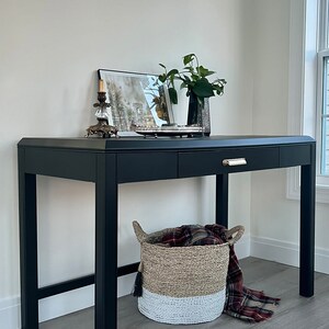 May include: Black desk with two drawers and a gold handle. The desk has a black painted finish and sits on a wood floor. A basket with a plaid blanket is in front of the desk.