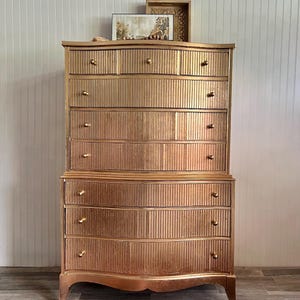May include: A tall, gold-painted wooden dresser with multiple drawers and a curved bottom. The dresser has a vertical grooved design and gold knobs. A framed picture and decorative tray sit on top. A patterned rug is in the foreground.