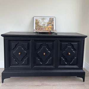 May include: A black wooden three-door cabinet with ornate carved details and brass knobs. The cabinet has a lower shelf and sits on four legs.