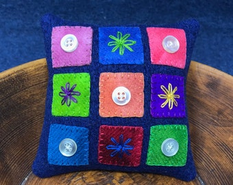 May include: A small square pillow with a patchwork design. The pillow is made of felt and features a variety of colors, including blue, green, red, orange, and pink. Each square has a different design, including buttons and embroidered flowers.