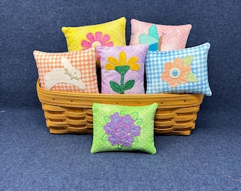 May include: Six small, square, fabric pillows with different embroidered designs. The pillows are in a wicker basket on a blue background. The designs include a butterfly, a flower, a bunny, and a flower with a button center.