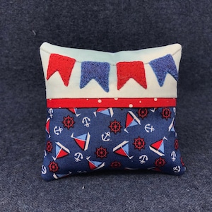 May include: A small, square pillow with a nautical theme. The front of the pillow is blue with white anchors and sailboats. The top of the pillow is white with red and blue bunting.