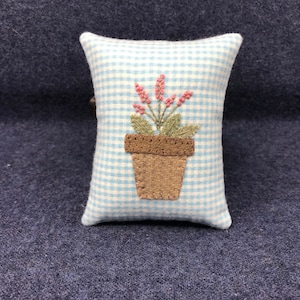 May include: A small blue and white gingham pillow with an embroidered image of a potted plant with pink flowers.