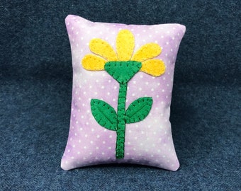 May include: A small purple pillow with white polka dots and a yellow flower with green leaves and stem appliqued on the front.
