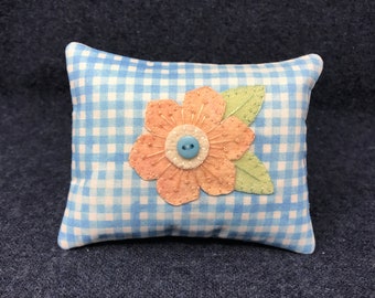 May include: A small, rectangular pillow with a blue and white gingham pattern. A peach-colored flower with a light blue button in the center is appliqued on the pillow.