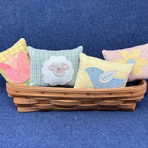 May include: A woven basket holds four decorative pillows. The pillows feature spring-themed designs: a pink tulip, a lamb, a blue bird, and a yellow flower. The pillows have various patterns and colors, including gingham and floral.