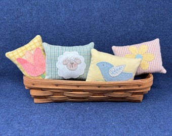 May include: A woven basket holds four decorative pillows. The pillows feature spring-themed designs: a pink tulip, a lamb, a blue bird, and a yellow flower. The pillows have various patterns and colors, including gingham and floral.