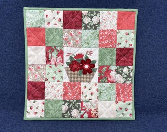 May include: A square quilted placemat with a patchwork design. The squares feature floral patterns in red, white, and green. A central square showcases a basket of red flowers. The placemat is trimmed with a green border.