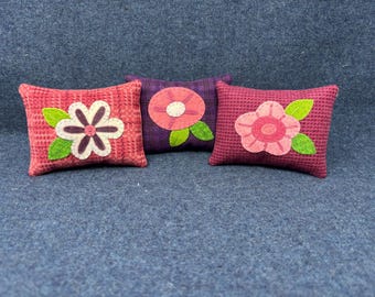 May include: Three decorative pillows with pink and green floral appliques. The pillows are made of a textured fabric and have a pink and purple color scheme.