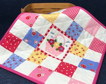 May include: A pink and white patchwork quilt with a floral embroidery design in the center. The quilt is made of various fabrics with different patterns, including polka dots, gingham, and floral prints.