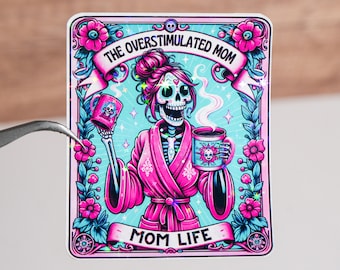 Puede incluir: Una pegatina con una ilustración rosa y azul de un esqueleto con una bata rosa y sosteniendo una taza de café. El texto "The Overstimulated Mom" está por encima del esqueleto y "Mom Life" está por debajo. La pegatina tiene un acabado brillante e iridiscente.