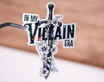 Könnte beinhalten: Ein holografischer Aufkleber mit einer Schwarzweißillustration eines Schwertes mit Blumen und dem Text "In My Villain Era".