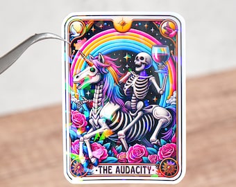 May include: A sticker featuring a skeleton riding a rainbow unicorn with a glass of wine in its hand. The sticker has a black background with stars and a rainbow arch. The text "THE AUDACITY" is printed at the bottom of the sticker.