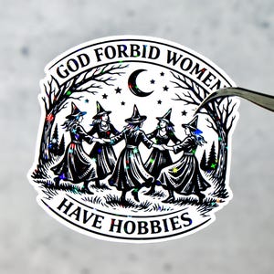 May include: A sticker with the text "GOD FORBID WOMEN HAVE HOBBIES" encircling a graphic of five witches dancing in a circle. The design is black and white with a crescent moon, stars, and trees.
