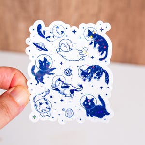 May include: A holographic sticker sheet featuring various cartoon cats in space helmets, floating among stars and planets. The cats are depicted in different poses and colors, with a whimsical and playful design.