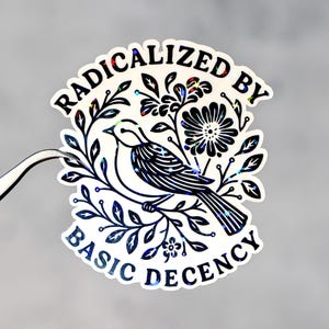 Könnte beinhalten: Ein holografischer Aufkleber mit einem schwarz-weißen Vogelmotiv, umgeben von floralen Elementen. Der Aufkleber trägt den Text "RADICALIZED BY BASIC DECENCY" in einem kreisförmigen Design. Eine silberne Pinzette hält den Aufkleber.