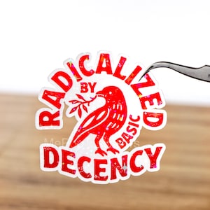 Op de afbeelding: Een sticker met de woorden "RADICALIZED BY BASIC DECENCY" in het rood, rondom een rode vogel die een takje vasthoudt. De sticker heeft een holografisch, glinsterend effect. Een pincet houdt de sticker vast.