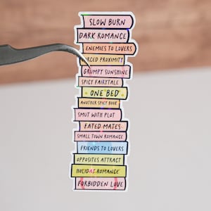May include: A stack of colorful stickers with different romance novel tropes written on them, including "Slow Burn", "Dark Romance", "Enemies to Lovers", "Forced Proximity", "Grumpy Sunshine", "Spicy Fairytale", "One Bed", "Another Spicy Book", "Smut with Plot", "Fated Mates", "Small Town Romance", "Friends to Lovers", "Opposites Attract", "Holiday Romance", and "Forbidden Love".