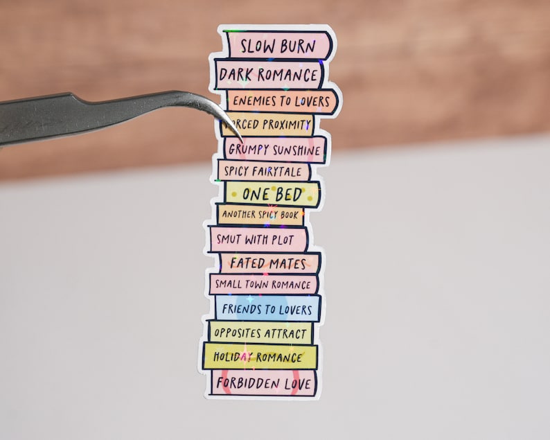 May include: A stack of colorful stickers with different romance novel tropes written on them, including "Slow Burn", "Dark Romance", "Enemies to Lovers", "Forced Proximity", "Grumpy Sunshine", "Spicy Fairytale", "One Bed", "Another Spicy Book", "Smut with Plot", "Fated Mates", "Small Town Romance", "Friends to Lovers", "Opposites Attract", "Holiday Romance", and "Forbidden Love".