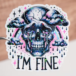 Puede incluir: Una pegatina holográfica con una calavera llorando lágrimas de arcoíris y nubes. El texto "I'M FINE" está debajo de la calavera.