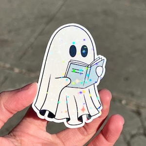 May include: A white ghost with a sparkly iridescent finish is reading a book. The ghost is wearing a white dress with a sparkly iridescent finish. The book is blue with a white cover and has a sparkly iridescent finish.