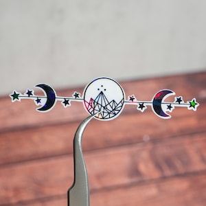 May include: A holographic sticker with a black and white design of a mountain with stars and moons. The sticker is being held by a pair of tweezers.