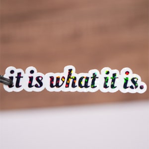 以下が含まれることがあります： 虹色のグリッターで飾られた黒い文字「it is what it is.」が書かれた白いステッカー