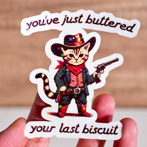 May include: A white sticker with a cartoon cat wearing a cowboy hat and holding a gun. The text on the sticker reads "you've just buttered your last biscuit".