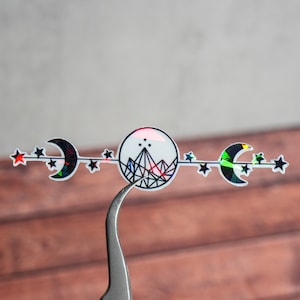 May include: A holographic sticker with a black and white design of a mountain with stars and crescent moons. The sticker has a rainbow iridescent finish.