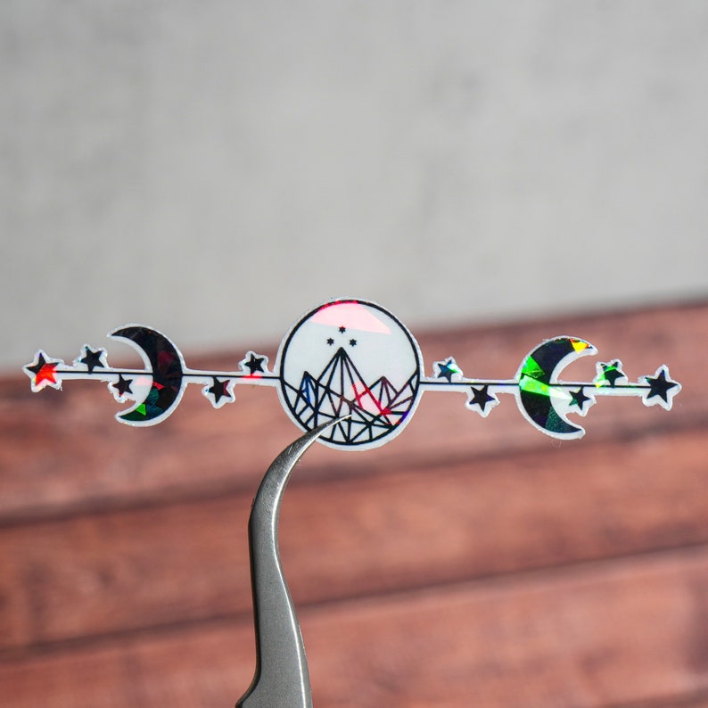 May include: A holographic sticker with a black and white design of a mountain with stars and crescent moons. The sticker has a rainbow iridescent finish.