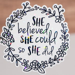 Può includere: Adesivo bianco con una ghirlanda di fiori neri e il testo "She believed she could so she did."