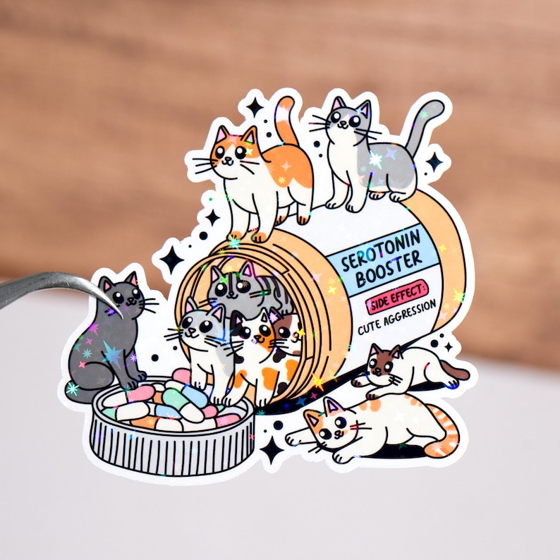 May include: A cartoon illustration of a pill bottle labeled "Serotonin Booster" with a side effect of "Cute Aggression". The bottle is filled with colorful pills and surrounded by adorable cartoon cats.
