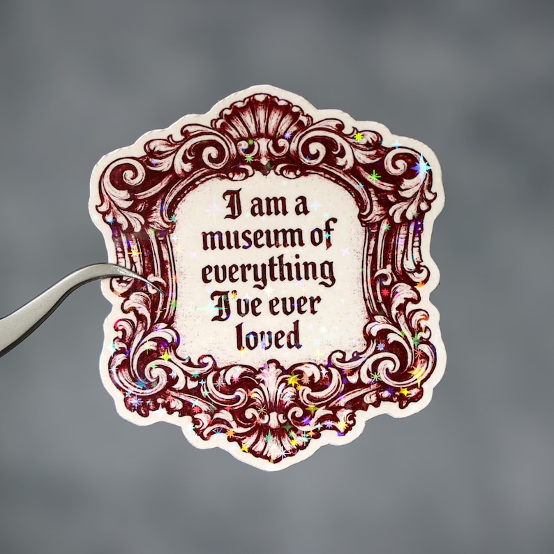 May include: A decorative sticker with a burgundy ornate frame and the text "I am a museum of everything I've ever loved." The sticker has a holographic, glittery effect. A pair of tweezers is holding the sticker.