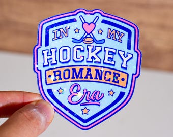 Może przedstawiać: Naklejka z błyszczącym niebieskim i różowym wzorem z tekstem "In My Hockey Romance Era" i różowym sercem z skrzyżowanymi kijami hokejowymi.