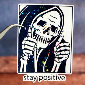 May include: A black and white sticker featuring a skeleton wearing a hooded robe and giving a thumbs up. The background is a glittery blue with white stars. The text "stay positive" is printed below the image.