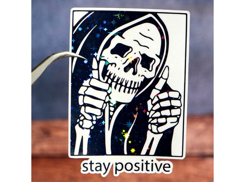 Puede incluir: Una pegatina en blanco y negro con un esqueleto que lleva una t&uacute;nica con capucha y hace un pulgar hacia arriba. El fondo es azul brillante con estrellas blancas. El texto "stay positive" est&aacute; impreso debajo de la imagen.