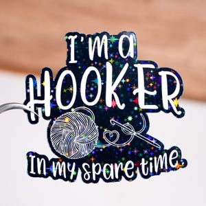May include: A black sticker with the text "I'm a HOOKER in my spare time" in white. The sticker features a yarn ball, crochet hook, and colorful star accents.