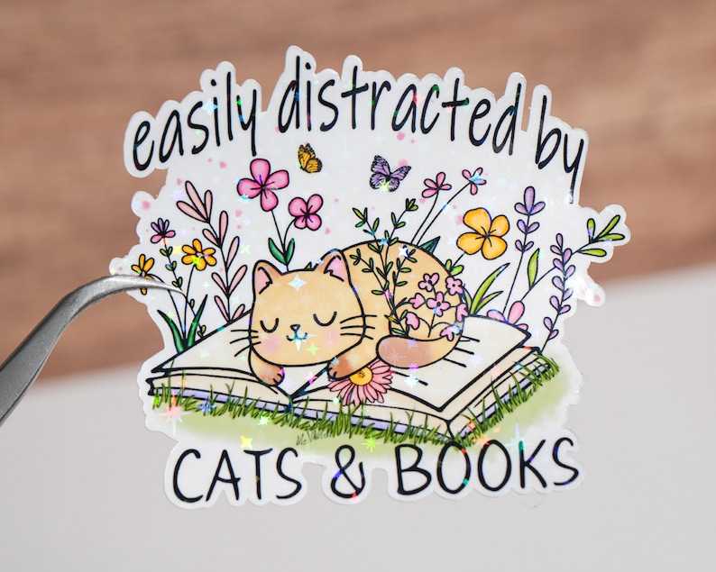May include: A sticker with a cat sleeping on a book surrounded by flowers and butterflies. The text reads "easily distracted by cats & books".