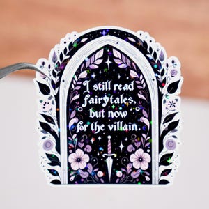 K&ouml;nnte beinhalten: Ein Aufkleber mit schwarzem Hintergrund und einem wei&szlig;en Bogenrahmen. Der Aufkleber tr&auml;gt den Text "I still read fairytales, but now for the villain." Das Design umfasst Sterne, Blumen und ein Schwert.