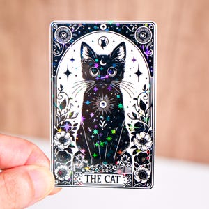 May include: A holographic sticker featuring a black cat tarot card design. The cat is surrounded by stars, moons, and floral elements, with the text "THE CAT" at the bottom. The sticker has a shiny, iridescent finish.