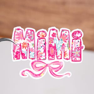 Puede incluir: Una pegatina rosa y floral con la palabra "Mimi" y un lazo a juego. La palabra "Mimi" está en una fuente en negrita, llena de una variedad de flores coloridas. El lazo está debajo de la palabra, con detalles de purpurina.