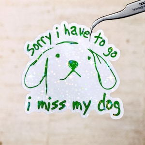 May include: A white sticker with a holographic sparkle effect, featuring a green outline of a dog's face and the text "Sorry I have to go, I miss my dog" in green. The sticker is being held by a pair of tweezers.