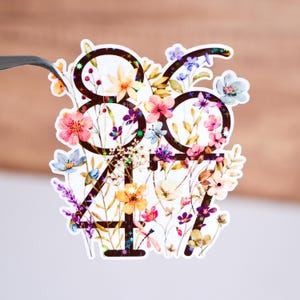 Peut inclure: Un autocollant avec les chiffres 8647 entourés de motifs floraux colorés. Les chiffres sont noirs et les fleurs sont dans des tons de rose, jaune, bleu et violet.