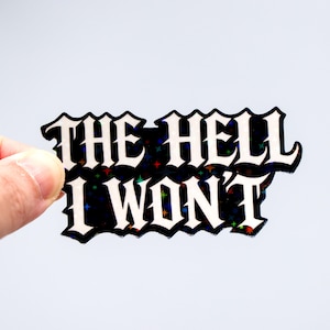 Op de afbeelding: Een zwarte sticker met de woorden "THE HELL I WON'T" in witte gotische letters. De sticker heeft een holografisch, glinsterend effect met kleurrijke sterren. De sticker wordt door een hand vastgehouden.