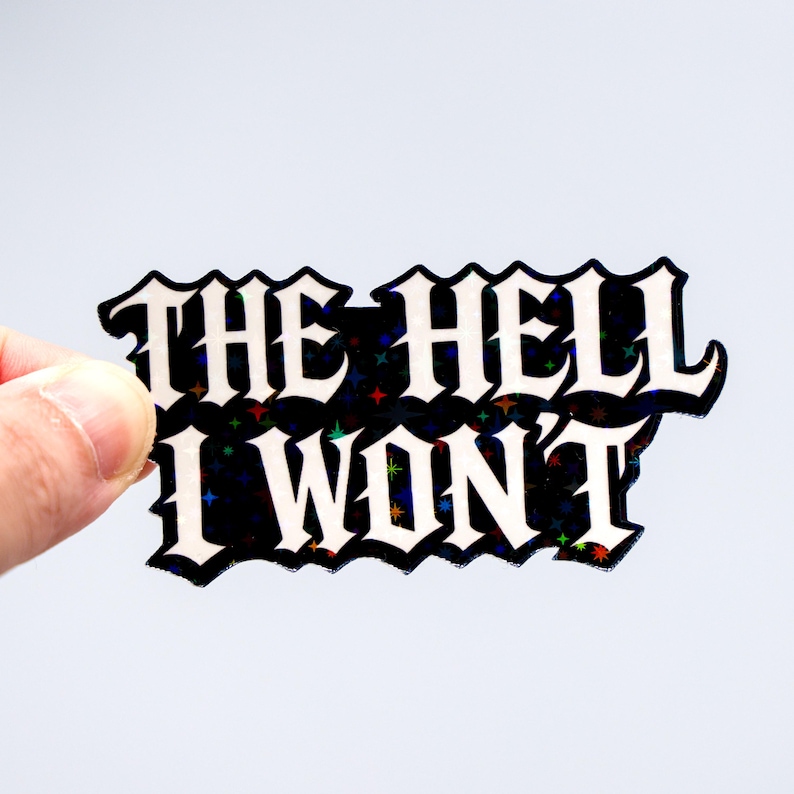 May include: A black sticker with the words "THE HELL I WON'T" in white gothic lettering. The sticker has a holographic, glittery effect with colorful stars. The sticker is held by a hand.